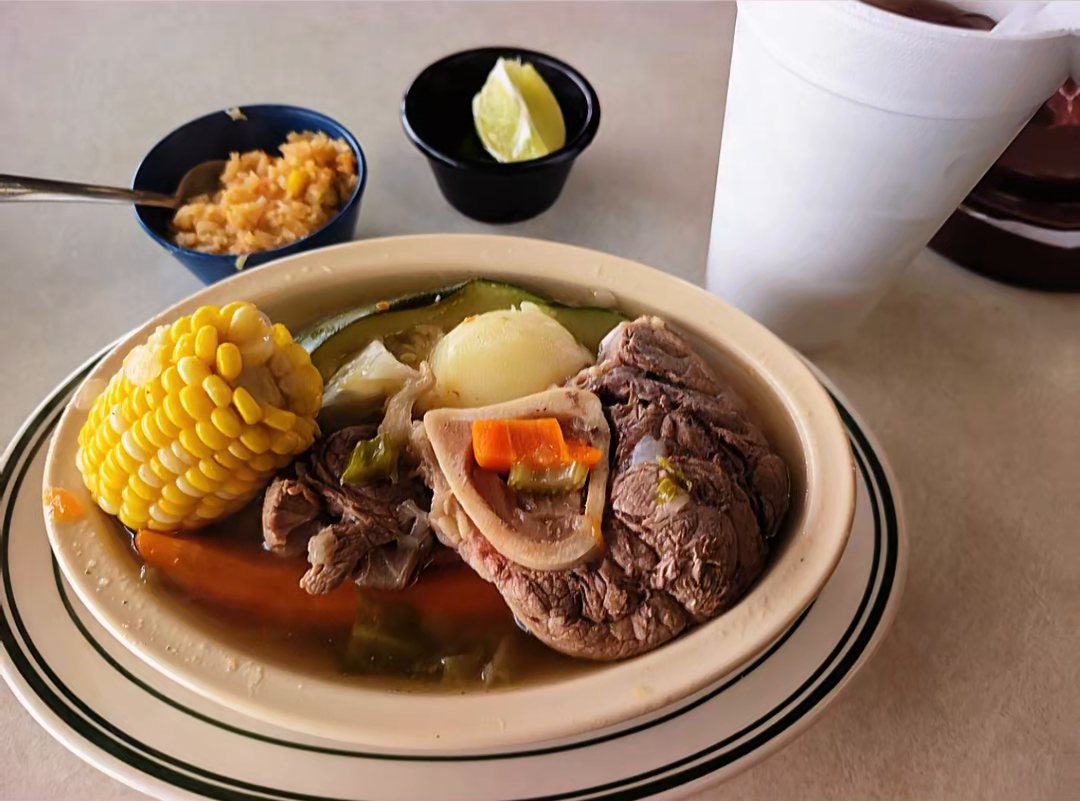 Large bowl of caldo de res with a beef shank on the bone, corn on the cob, potato, zucchini and carrots, with rice and lime on the side